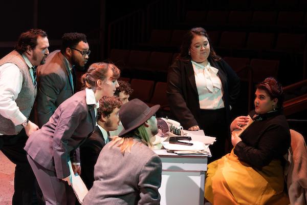 Students performing in 9 to 5, gathered around someone at a desk